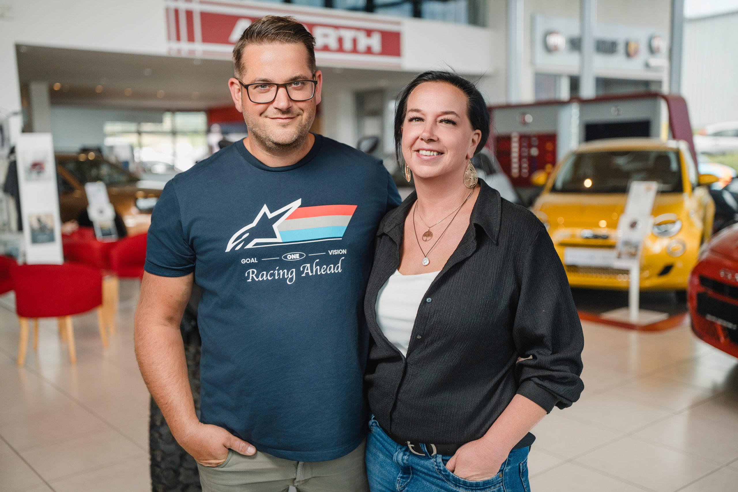 Zwei Mitarbeiter stehen lächelnd im Autohaus vor ausgestellten Fahrzeugen