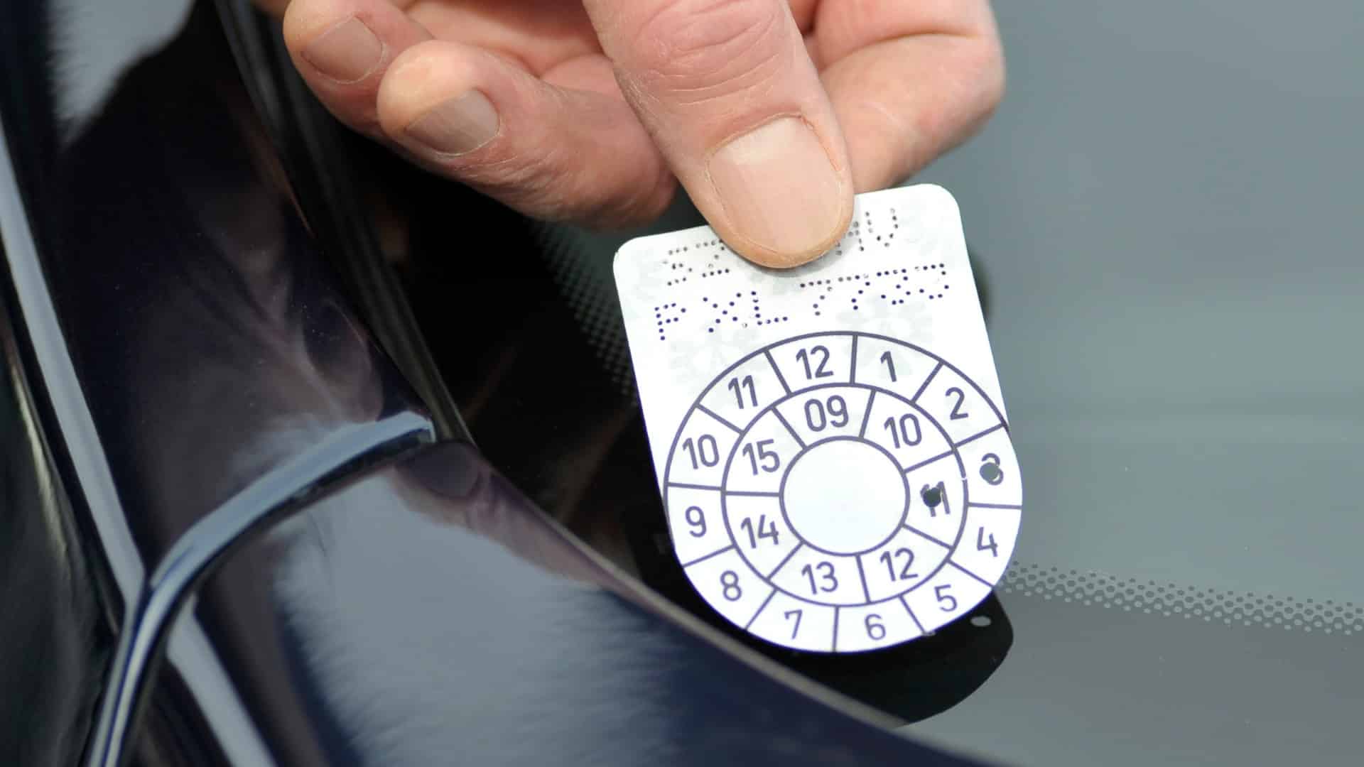 Eine Hand bringt eine § 57a-Begutachtungsplakette an der Windschutzscheibe eines Autos an.